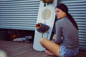 Brunette girl in gray turtleneck and denim shorts is posing with her wakeboard sitting on a floor near an equipment storage garage. Close-up. photo