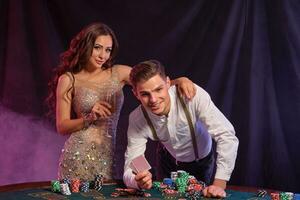 Man and woman playing poker at casino, celebrating win at table with stacks of chips, cards, champagne. Black, smoke background. Close-up. photo