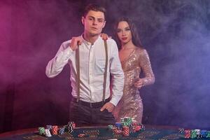 Man and woman playing poker at casino, celebrating win at table with stacks of chips, cards, champagne. Black, smoke background. Close-up. photo