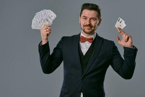 Man in black classic suit and red bow-tie showing two playing cards and cash, posing on gray studio background. Gambling, poker, casino. Close-up. photo