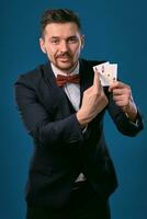 Man in black classic suit and red bow-tie showing two playing cards while posing against blue studio background. Gambling, poker, casino. Close-up. photo