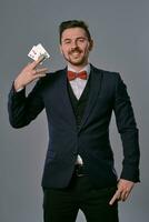 Man in black classic suit and red bow-tie showing two playing cards and chips, posing on gray studio background. Gambling, poker, casino. Close-up. photo