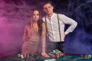 hombre y mujer jugando póker a casino, celebrando ganar a mesa con pilas de papas fritas, tarjetas, champán. negro, fumar antecedentes. de cerca. foto