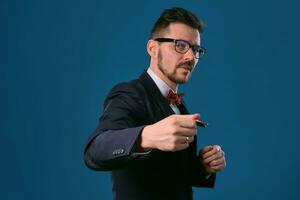 Man in black classic suit, red bow-tie, glases is showing two colored chips, posing on blue studio background. Gambling, poker, casino. Close-up. photo