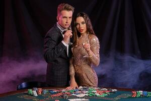 Man and woman playing poker at casino, celebrating win at table with stacks of chips, money, cards, champagne. Black, smoke background. Close-up. photo