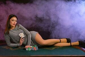 Girl in gray dress playing poker at casino, holding cards, laying on table with chips on it. Black, smoke background. Gambling. Close-up. photo