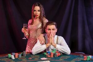 Man playing poker at casino sitting at table with stacks of chips, money, champagne, cards. Celebrating win with woman. Black background. Close-up. photo