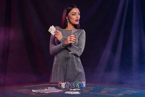 Girl playing poker at casino, holding champagne, two aces. Posing at table with chips, money, cards on it. Black, smoke background. Close-up. photo