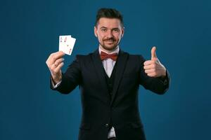 Man in black classic suit and red bow-tie showing two playing cards while posing against blue studio background. Gambling, poker, casino. Close-up. photo