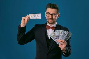 Man in black classic suit, red bow-tie, glases is holding some dollar bills, posing on gray studio background. Gambling, poker, casino. Close-up. photo