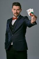 Man in black classic suit and red bow-tie showing two playing cards and chips, posing on gray studio background. Gambling, poker, casino. Close-up. photo