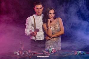 Man and woman playing poker at casino, celebrating win at table with stacks of chips, cards, champagne. Black, smoke background. Close-up. photo