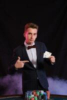 Elegant man is playing poker at casino. He is posing at the table with stacks of colored chips on it, against a black, smoke background. photo