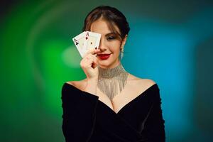 Brunette girl in black dress, necklace and earrings. Smiling, covered her eye by two aces, posing on colorful background. Poker, casino. Close-up photo