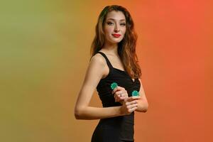 Brunette model with earring in nose, in black dress. Holding two green chips, posing sideways on colorful background. Poker, casino. Close-up photo