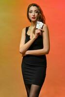 Brunette girl with earring in nose, in black dress. Showing two aces while posing on colorful background. Gambling, poker, casino. Close-up photo