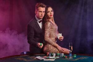 Man and woman playing poker at casino, celebrating win at table with stacks of chips, money, cards, champagne. Black, smoke background. Close-up. photo