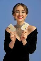 Brunette model with bare shoulders, in black dress and jewelry. Smiling, showing two playing cards posing on blue background. Poker, casino. Close-up photo