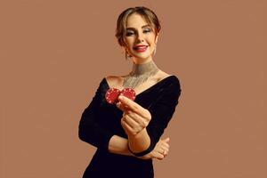 Brunette female in black dress and shiny jewelry. She smiling, showing two red chips, posing on brown background. Poker, casino. Close-up photo