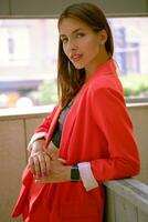 Blonde girl in red lady-type pantsuit and black top, watch, ring, pendant around her neck is posing leaning on a wooden partition. Close-up. photo