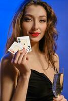 Brunette model with earring in nose, in black dress. Holding glass of champagne, two aces, smiling, posing on blue background. Poker, casino. Close-up photo