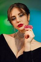 Cute brunette lady in black dress, necklace and earrings. She is showing two red chips, posing on colorful studio background. Poker, casino. Close-up photo