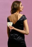 Brunette female in black dress. Showing two playing cards, posing standing back to camera against pink background. Poker, casino. Close-up photo