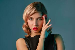 Blonde girl in black leather dress holding one red chip, posing against blue background. Gambling entertainment, poker, casino. Close-up. photo