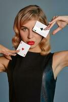 Blonde girl in black leather dress showing two playing cards, posing against gray background. Gambling entertainment, poker, casino. Close-up. photo