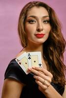 morena niña con arete en nariz, en negro vestido. demostración dos ases mientras posando en rosado estudio antecedentes. póker, casino. de cerca, Copiar espacio foto