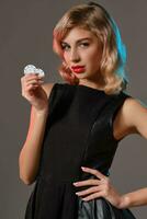 Blonde girl in black stylish dress holding two chips, posing against gray background. Gambling entertainment, poker, casino. Close-up. photo