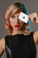 Blonde girl in black leather dress showing ace of clubs, posing against gray background. Gambling entertainment, poker, casino. Close-up. photo