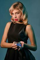 Blonde girl in black leather dress holding some colorful chips, posing against blue background. Gambling entertainment, poker, casino. Close-up. photo