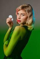 Blonde girl in black stylish dress holding two chips, posing against gray background, green flare. Gambling entertainment, poker, casino. Close-up. photo