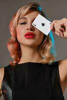 Blonde girl in black leather dress showing ace of clubs, posing against gray background. Gambling entertainment, poker, casino. Close-up. photo