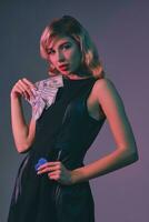 Blonde girl in black stylish dress holding some money and chips, posing against colorful background. Gambling entertainment, poker, casino. Close-up. photo