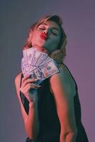 Blonde girl in black stylish dress holding some money, posing against colorful background. Gambling entertainment, poker, casino. Close-up. photo