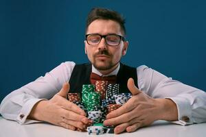 Man in glasses, black vest and shirt sitting at white table with stacks of chips on it, posing on blue background. Gambling, poker, casino. Close-up. photo