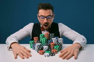 Guy in glasses, black vest and shirt sitting at white table with stacks of chips on it, posing on blue background. Gambling, poker, casino. Close-up. photo