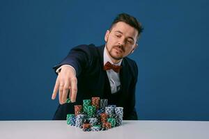 Man in black suit is sitting at white table with colored stacks of chips on it, posing on blue studio background. Gambling, poker, casino. Close-up. photo