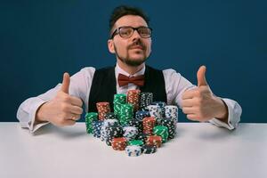 Player in glasses, black vest and shirt sitting at white table with stacks of chips on it, posing on blue background. Poker, casino. Close-up. photo