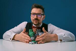 Male in glasses, black vest and shirt sitting at white table with stacks of chips on it, posing on blue background. Gambling, poker, casino. Close-up. photo