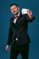 Man in black classic suit and red bow-tie showing two playing cards while posing against blue studio background. Gambling, poker, casino. Close-up. photo