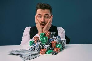 Man in black vest and shirt sitting at white table with stacks of chips and cash on it, posing on blue background. Gambling, poker, casino. Close-up. photo