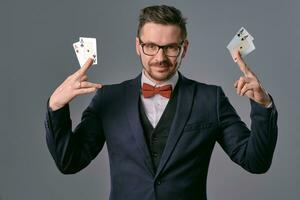 Man in black classic suit, red bow-tie, glases is showing four playing cards, posing on gray studio background. Gambling, poker, casino. Close-up. photo