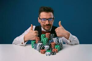 Fellow in glasses, black vest and shirt sitting at white table with stacks of chips on it, posing on blue background. Poker, casino. Close-up. photo