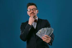 Man in black classic suit, red bow-tie, glases is holding some dollar bills, posing on gray studio background. Gambling, poker, casino. Close-up. photo