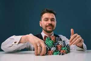 Man in black vest and shirt sitting at white table with stacks of chips on it, posing on blue studio background. Gambling, poker, casino. Close-up. photo