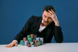 hombre en negro traje es sentado a blanco mesa con de colores pilas de papas fritas en él, posando en azul estudio antecedentes. juego, póker, casino. de cerca. foto