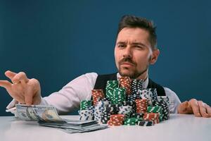 Man in black vest and shirt sitting at white table with stacks of chips and cash on it, posing on blue background. Gambling, poker, casino. Close-up. photo
